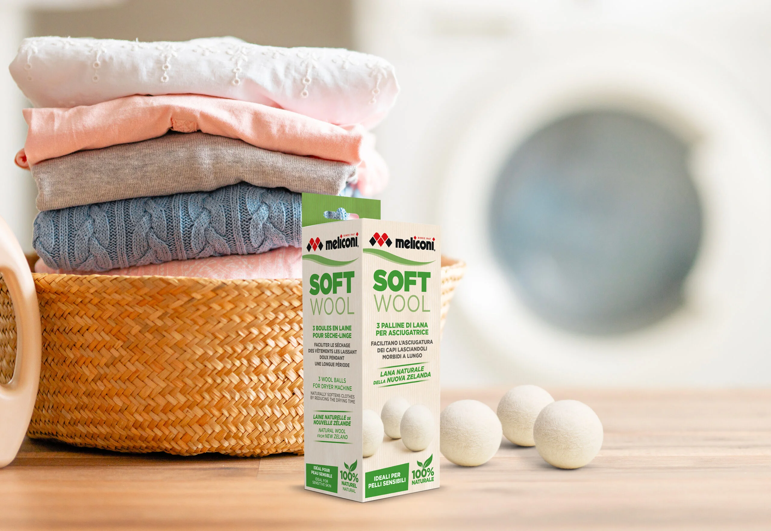 Balenie Meliconi Soft Wool a biele vlnené gule do sušičky na stole vedľa prúteného koša s poskladanou bielizňou.