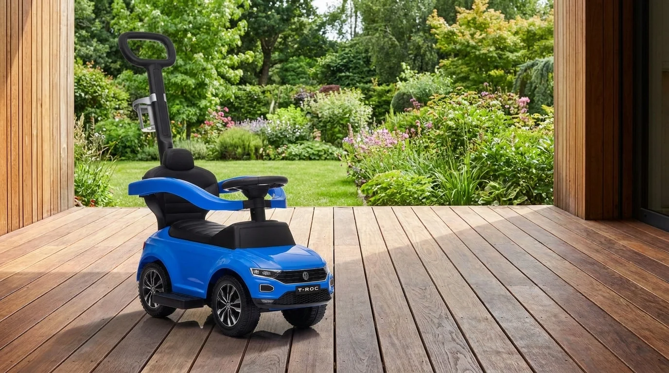 Modré detské odrážadlo Buddy Toys VW T-ROC s vodiacou tyčou stojí na drevenej terase pred zelenou záhradou.