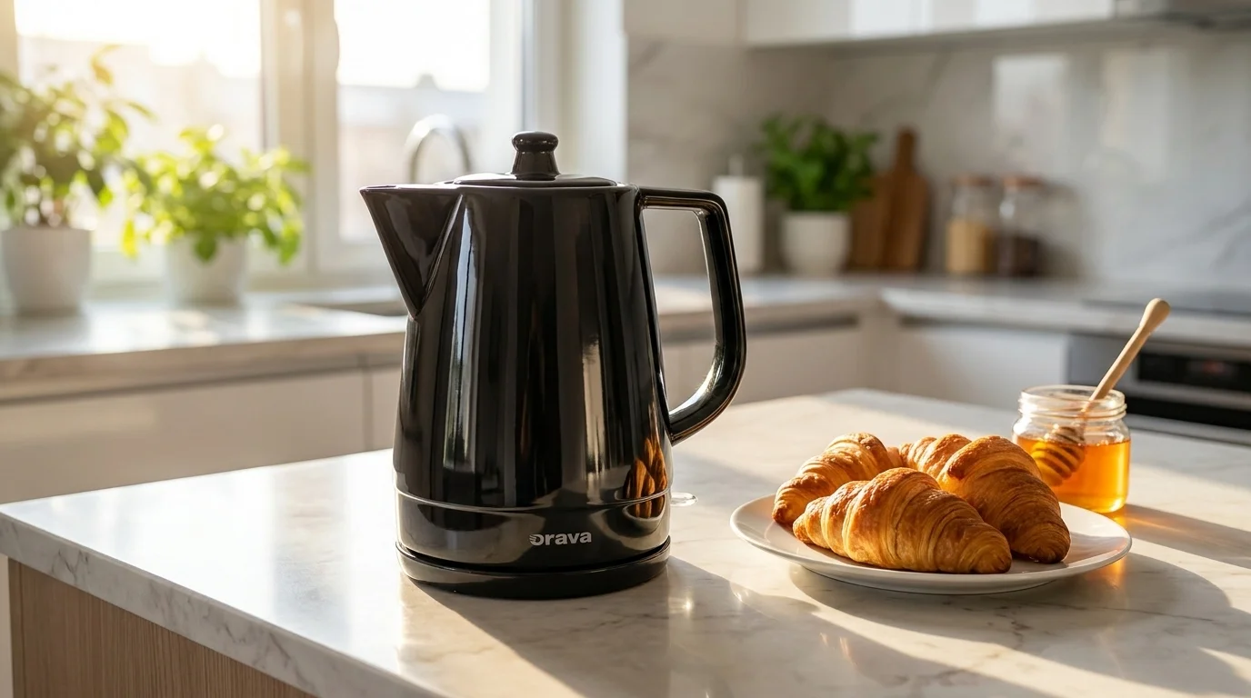 Černá keramická varná konvice Orava VK-3813 B stojí na kuchyňské lince vedle talíře s croissanty a sklenice s medem.