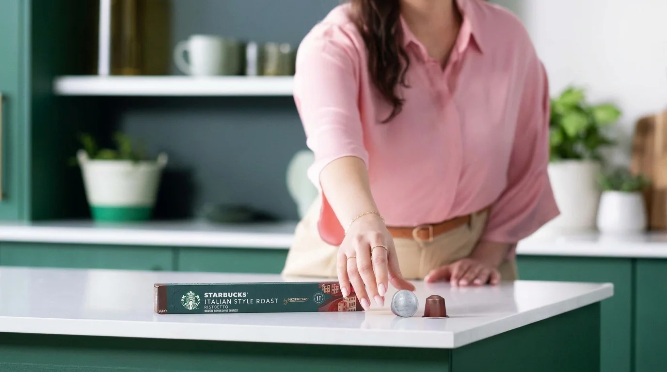 Žena v růžové košili sahá po kávové kapsli Starbucks Italian Style Roast na bílé kuchyňské lince.