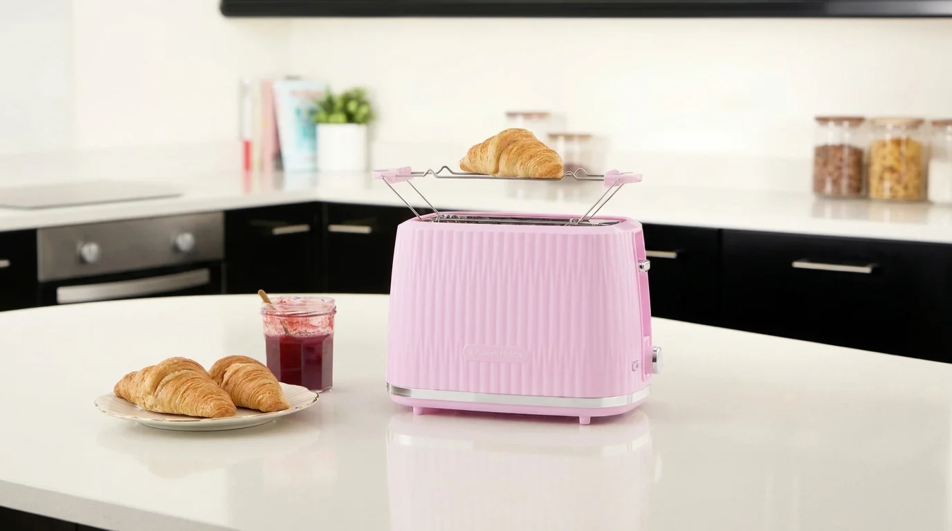 Růžový topinkovač Russell Hobbs s mřížkou na rozpékání pečiva stojí na bílé kuchyňské lince vedle talíře s croissanty a sklenice s džemem.