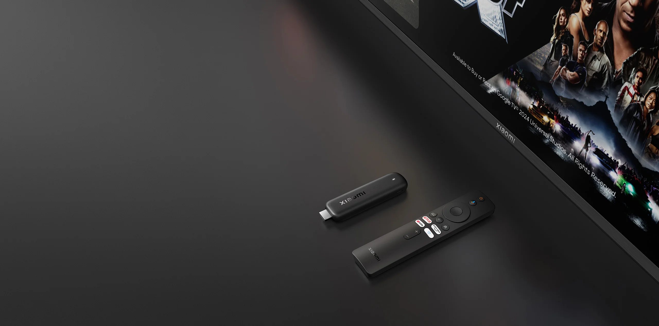 Černý Xiaomi TV Stick HD a dálkový ovladač ležící na tmavém povrchu před televizní obrazovkou.