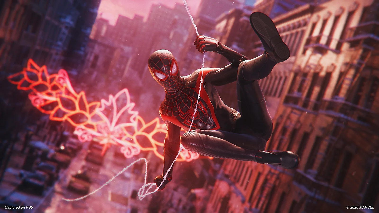 Miles Morales v čierno-červenom obleku Spider-Mana sa hojdá na pavučine nad mestskou ulicou so svietiacimi ozdobami v pozadí.