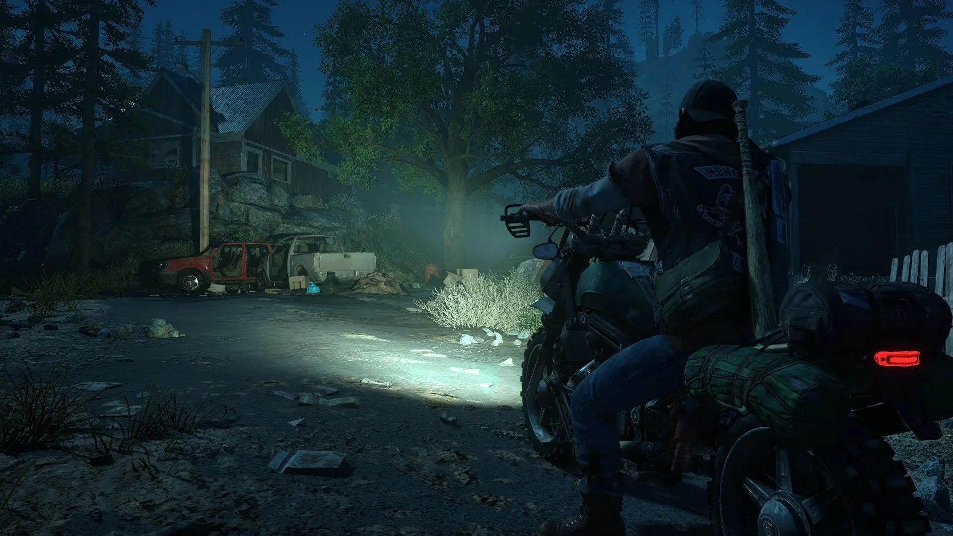 Deacon St. John na motorce s rozsvíceným světlometem projíždí v noci kolem opuštěných aut a chaty v lese ve hře Days Gone.
