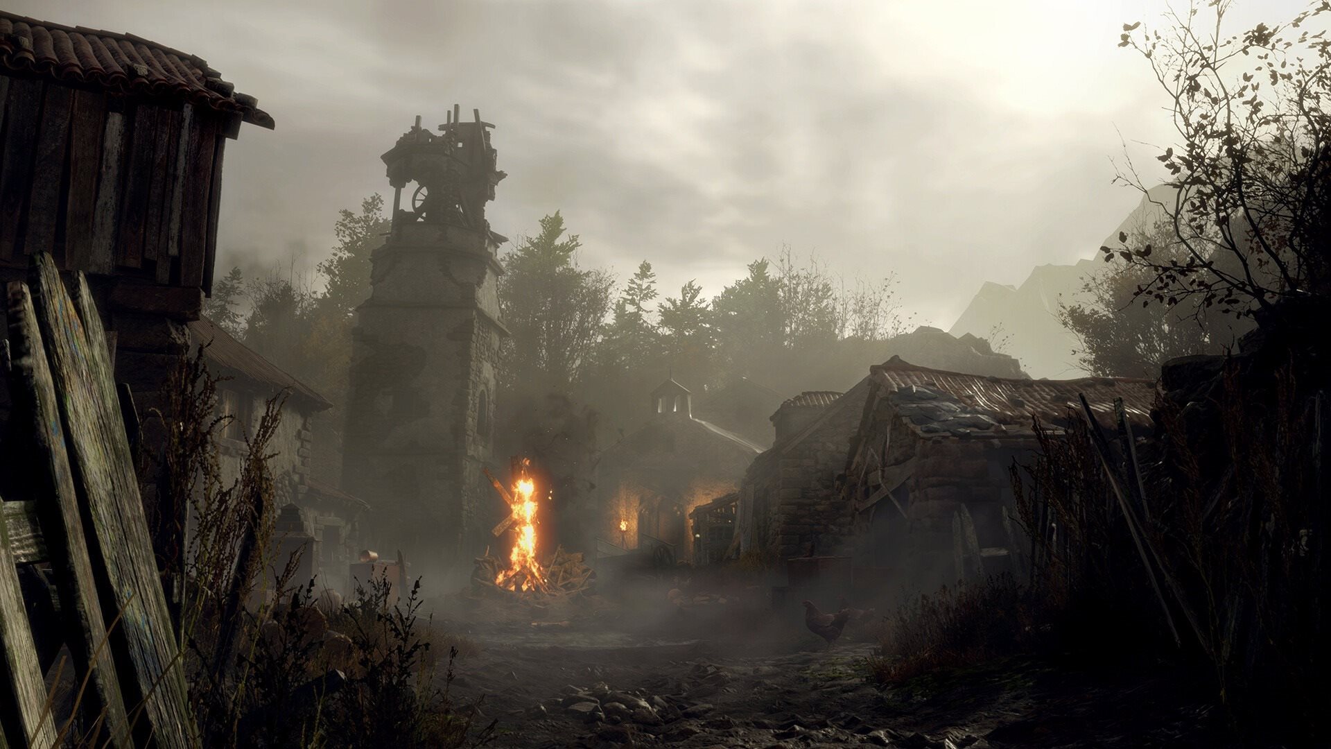 Temná a mlhavá vesnice ze hry Resident Evil 4 s hořícím ohništěm a kamennou věží pod zataženou oblohou.