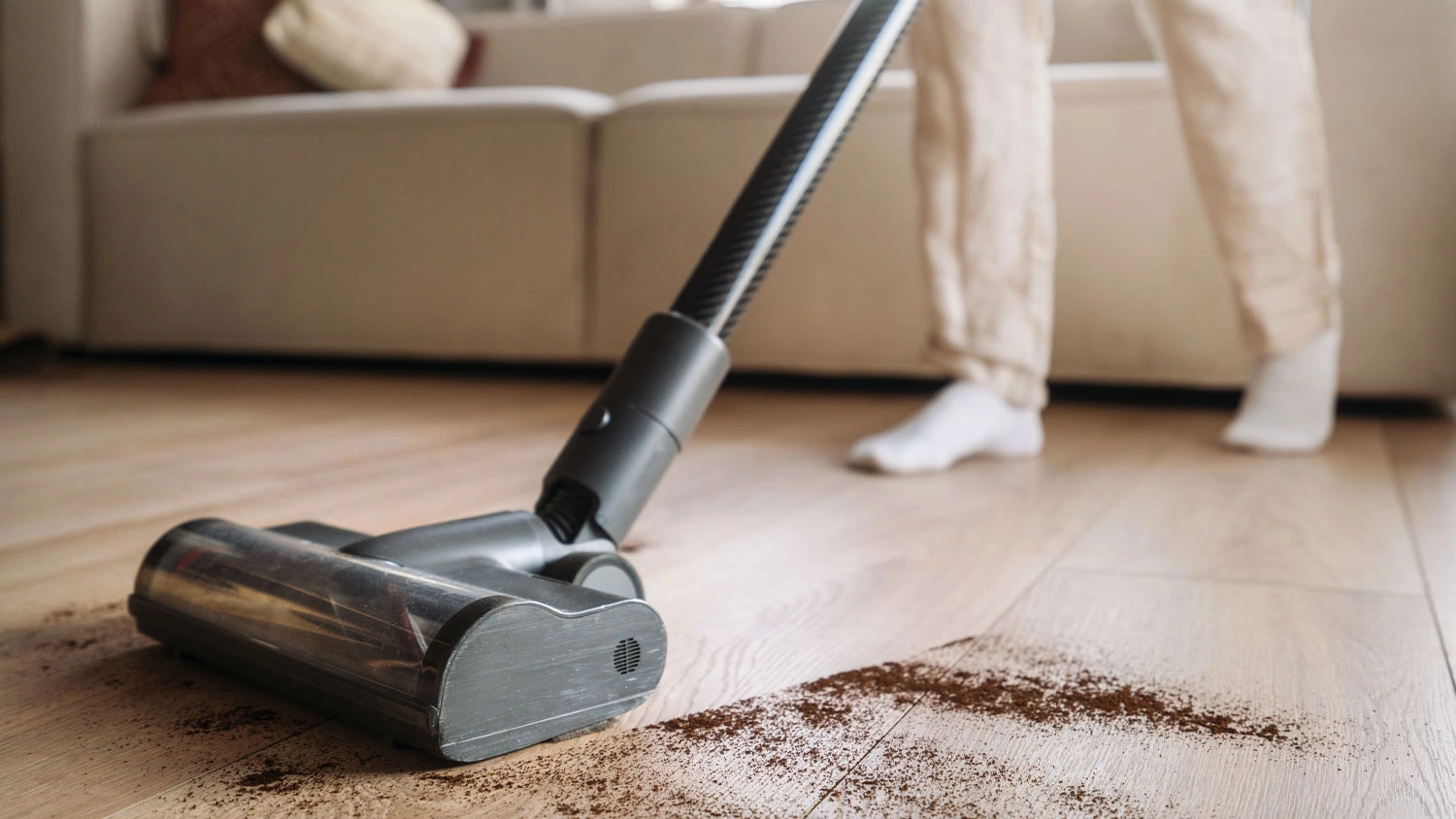 Tyčový vysavač Hoover vysává hnědé nečistoty ze světlé dřevěné podlahy.