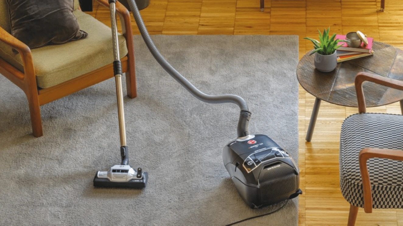 Černý vysavač Hoover při vysávání šedého koberce v obývacím pokoji s dřevěnou podlahou.