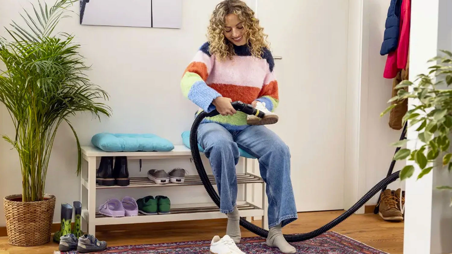 Žena sedí na lavičke v predsieni a čistí bielu tenisku pomocou nadstavca na čistenie topánok Kärcher Shoe!Cleaner.