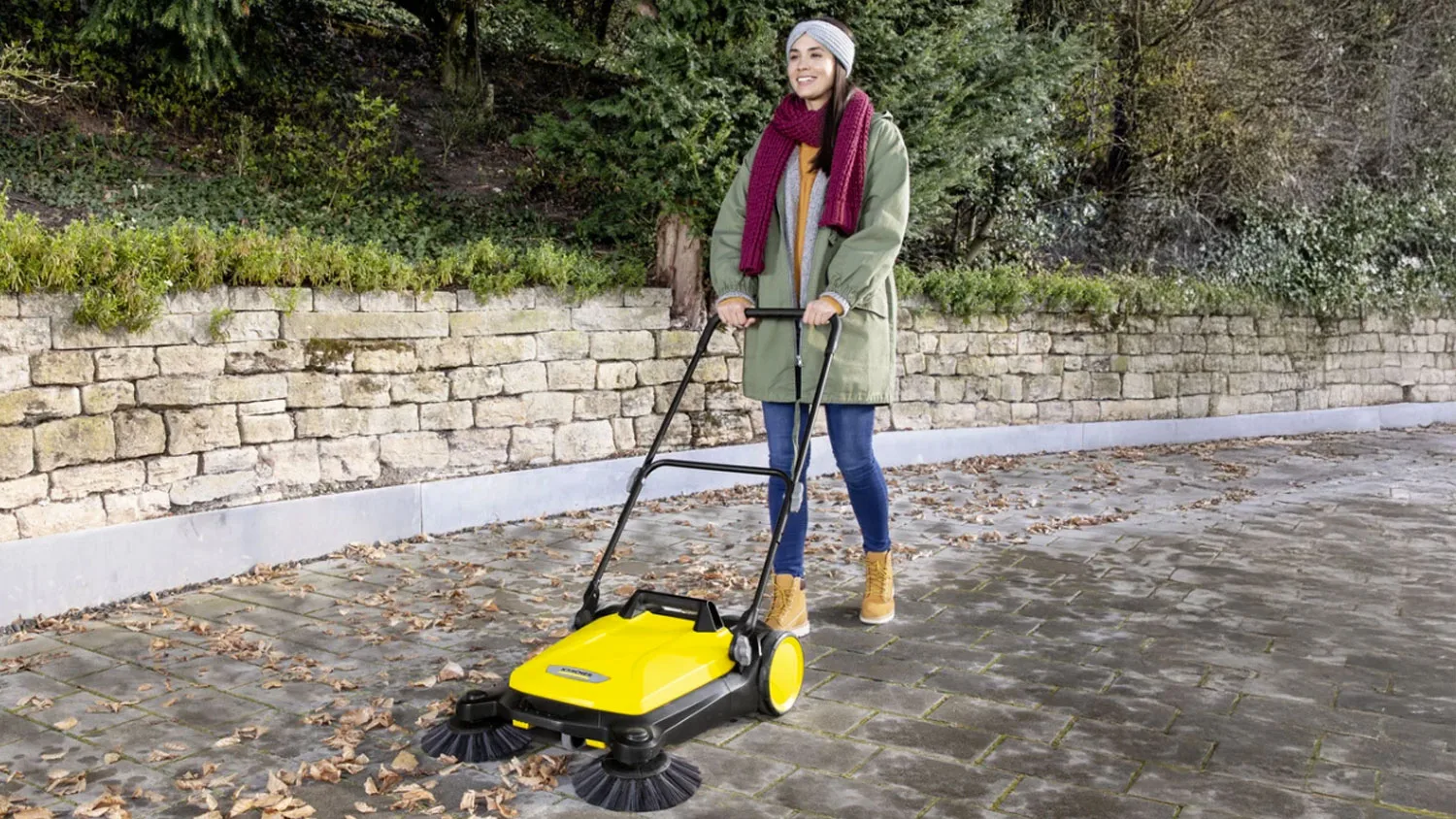 Žena na dlážděném chodníku zametá spadané listí pomocí žlutého zametacího stroje Kärcher s bočními kartáči.