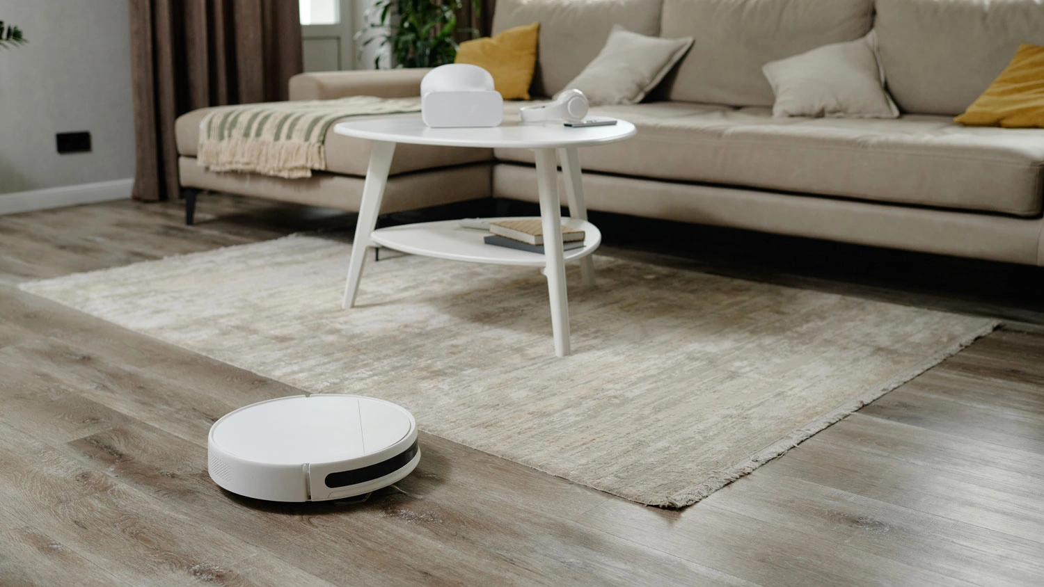 Bílý robotický vysavač Sencor na dřevěné podlaze v obývacím pokoji vedle světlého koberce a konferenčního stolku.