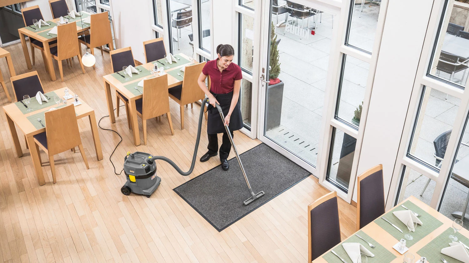 Žena v restauraci vysává rohožku u dveří pomocí mokro-suchého vysavače Kärcher NT 27/1.