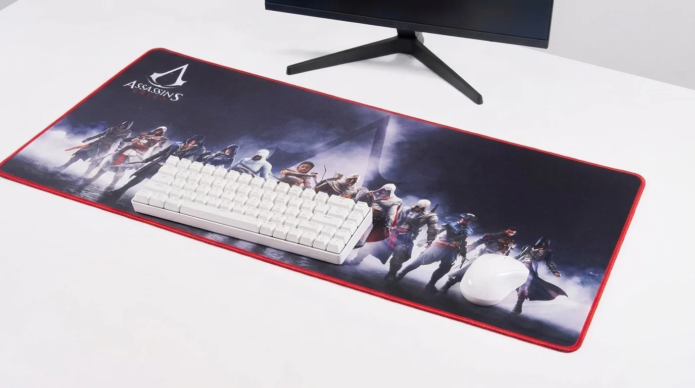 Velká herní podložka Superdrive Assassin's Creed XXL na stole s položenou bílou klávesnicí a myší pod monitorem.