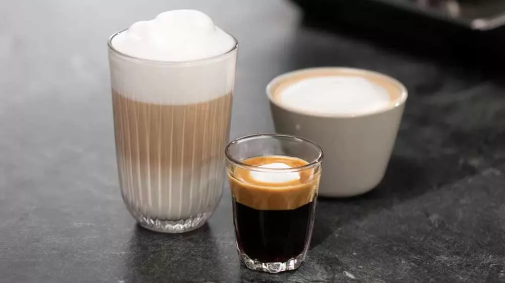 Vysoký pohár s vrstveným latte macchiato, malý pohár s espressom a šálka kávy s mliečnou penou na tmavej ploche.