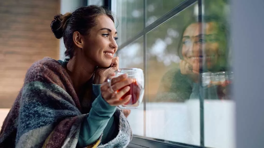 Usmievavá žena zabalená v deke sa pozerá z okna a v rukách drží sklenenú šálku s horúcim čajom.