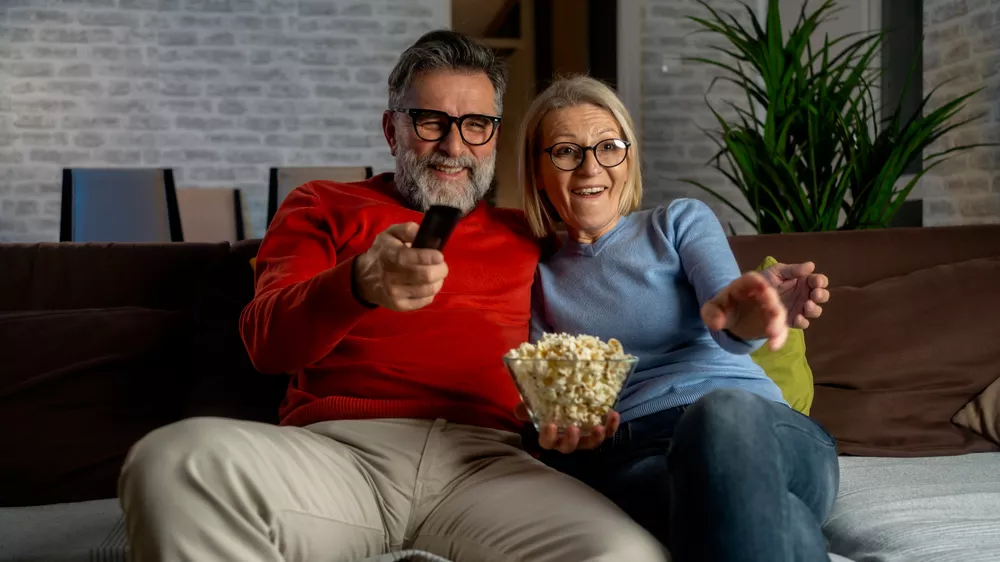 Usmiaty starší pár sedí na pohovke s miskou pukancov a muž mieri diaľkovým ovládačom dopredu.
