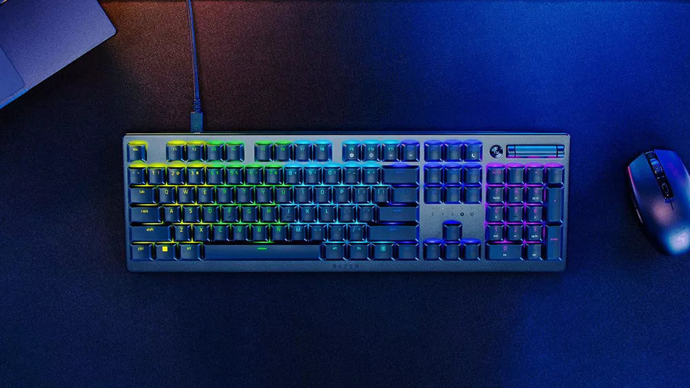 Herná klávesnica Razer s farebným RGB podsvietením položená na tmavom stole vedľa počítačovej myši.