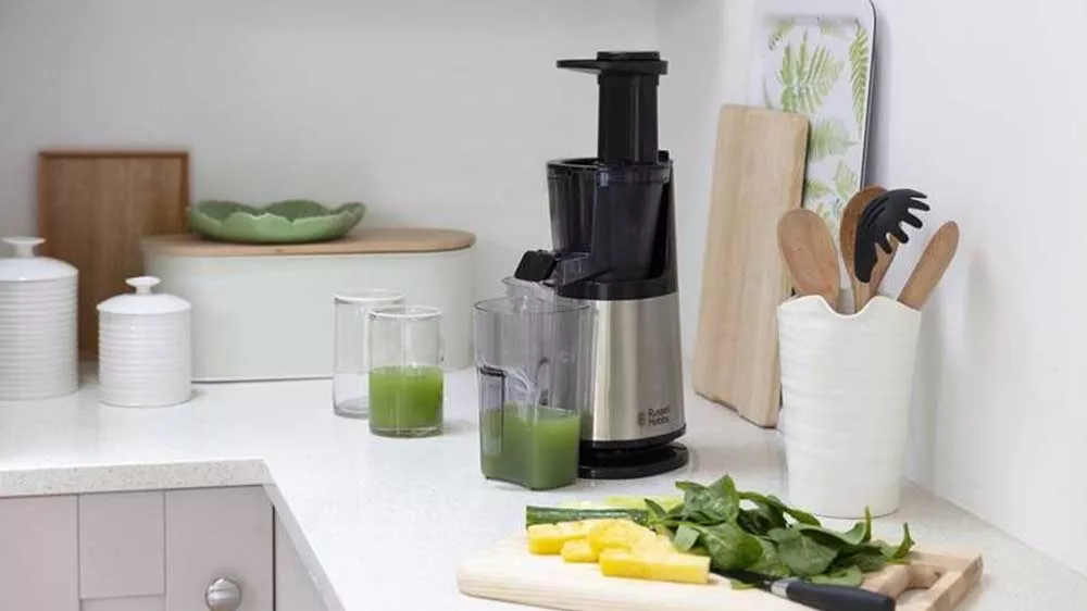 Pomalý odšťavovač Russell Hobbs na kuchynskej linke s nádobou so zelenou šťavou a nakrájaným ovocím a zeleninou na doske.