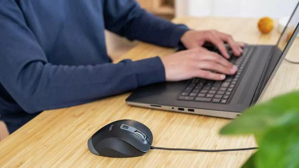 Čierna ergonomická myš Trust Fyda Eco na drevenom stole vedľa notebooku, na ktorom píše používateľ.